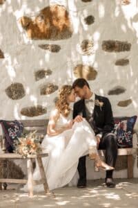 Braut und Bräutigam sitzen eng beieinander auf einer rustikalen Bank im gesprenkelten Sonnenlicht und verkörpern die perfekte Essenz des Heiratens. Die Braut in einem weißen Kleid und einem Blumenkopfschmuck ergänzt den dunklen Anzug des Bräutigams. In der Nähe verleiht ein Blumenstrauß dem Hintergrund aus Stein und weiß getünchten Wänden Charme.