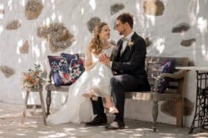Braut und Bräutigam sitzen auf einer Holzbank im Freien, umgeben von Steinmauern – eine perfekte Szene für eine intime Hochzeit. Die Braut im weißen Kleid hält die Hände des Bräutigams im schwarzen Anzug. Üppige Blumenkissen und ein Blumenstrauß bereichern die Kulisse, und das gesprenkelte Sonnenlicht wirft sanfte Schatten.