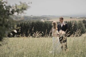 Auf einer malerischen Wiese stehen sich Braut und Bräutigam an ihrem Hochzeitstag liebevoll gegenüber. Die Braut in ihrem weißen Kleid mit einem Blumenstrauß in der Hand blickt ihren Bräutigam im Anzug an. Hinter ihnen erstrecken sich sanfte Hügel und ein Waldgebiet unter dem klaren Himmel – eine perfekte Szene aus ihren Hochzeitsplanungsträumen.