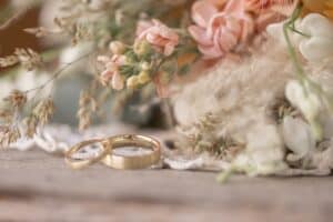 Zwei goldene Eheringe auf einer Holzoberfläche, umgeben von einem zarten Arrangement aus pastellfarbenen Blumen und zartem Grün, fangen die Essenz einer romantischen und rustikalen Hochzeit perfekt ein. Die Szene strahlt Liebe und Charme aus, ideal für jedes Paar, das davon träumt, in solch einer zauberhaften Umgebung zu heiraten.
