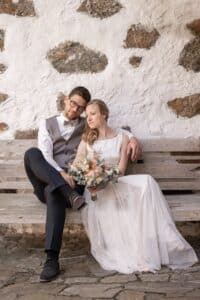 Ein Paar sitzt in eleganter Hochzeitskleidung auf einer rustikalen Holzbank und verkörpert einen wunderschönen Hochzeitsmoment. Der Bräutigam in grauer Weste und weißem Hemd und die Braut in ihrem weißen Kleid mit einem Blumenstrauß in der Hand lehnen an einer alten Steinmauer und strahlen Liebe und Ruhe aus.