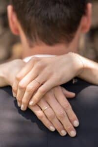 Eine Nahaufnahme eines sich umarmenden Paares fängt einen Moment zärtlicher Intimität ein. Die Arme der Frau, geschmückt mit einem kleinen Armband und einem einfachen Ring, schlingen sich um die Schultern des Mannes. Ihre goldenen Eheringe, Symbole der Liebe und Heiraten, glänzen sanft in dieser gefühlvollen Szene.