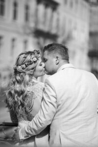 Ein Schwarzweißfoto fängt einen zärtlichen Kuss zwischen einem Paar ein, das seinen Hochzeitsmoment im Freien genießt. Die Frau, geschmückt mit einem Blumenstirnband und sichtbarem Schultertattoo, lehnt sich an den Mann im hellen Anzug. Sie teilen diesen intimen Moment in einer städtischen Umgebung mit verschwommenen Gebäuden im Hintergrund.