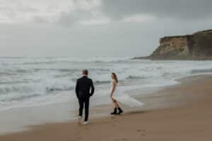 Ein Paar, das vielleicht über seine Hochzeitsplanung nachdenkt, spaziert an einem Sandstrand entlang, in dessen Nähe die Wellen brechen. Die Frau trägt ein fließendes weißes Kleid und schwarze Stiefel, während der Mann einen schwarzen Anzug trägt. Der Himmel ist bedeckt und im Hintergrund ragt eine Klippe auf, die ihrem Moment einen Hauch Dramatik verleiht.