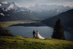 Braut und Bräutigam schwelgen in ihrem Hochzeitsglück und gehen Händchen haltend einen grasbewachsenen Hügel entlang, von dem aus man einen See und schneebedeckte Berge überblicken kann. Der Himmel ist klar und die Szene ist ruhig und malerisch.
