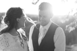 Ein Schwarzweißfoto zeigt ein Paar, das sich im Freien anlächelt und die Freude einer Hochzeit verkörpert. Die Frau mit ihrem langen Haar, das mit einem Blumenschmuck geschmückt ist, trägt ein Spitzenkleid. Der Mann in Weste und Krawatte steht neben ihr, während das Sonnenlicht einen sanften Lichtschein um sie herum erzeugt.