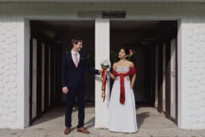 Ein Paar steht unter einem Torbogen und verkörpert die Essenz einer Hochzeit. Der Mann in seinem dunklen Anzug mit rosa Krawatte und die Frau in ihrem weißen, mit bunten Stickereien verzierten Kleid und einem roten Schal halten sich an den Händen. Sie hält einen Blumenstrauß, während sie einander in die Augen blicken und die Freude des Heiratens einfangen.