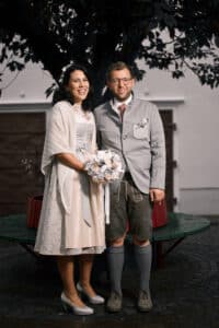 Ein Paar in Hochzeitskleidung, die Frau in einem beige-weißen Kleid und der Mann in traditioneller bayerischer Kleidung, steht lächelnd unter einem Baum. Die Braut hält einen Blumenstrauß in der Hand, der die Freude ihrer Hochzeit einfängt.