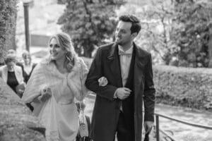 Braut und Bräutigam gehen Arm in Arm im Freien und verkörpern die Freude am Heiraten. Die Braut trägt ein weißes Kleid mit Schal und der Bräutigam einen Anzug mit Fliege. Bäume säumen ihren Weg und deuten auf eine sorgfältige Hochzeitsplanung hin, während die Gäste ihnen durch diese bezaubernde Szene folgen.
