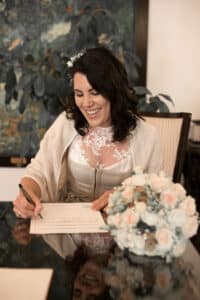 Eine Frau in weißem Kleid und Schal lächelt, während sie an einem Tisch ein Dokument unterschreibt, ganz in der Freude der Hochzeit versunken. Ein Strauß heller Blumen verleiht dieser formellen Hochzeitsplanung Eleganz.