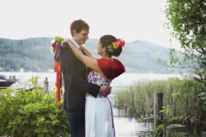 Ein Paar umarmt sich während seiner Hochzeit an einem ruhigen Seeufer. Die Frau trägt traditionelle, farbenfrohe Kleidung mit Blumen im Haar und der Mann trägt einen Anzug. Üppiges Grün und Berge bilden den Hintergrund unter einem teilweise bewölkten Himmel und schaffen eine idyllische Szene für Heiraten.