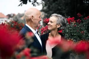 Ein älteres Paar steht eng beieinander und lächelt sich inmitten eines Gartens mit leuchtend roten Rosen an, was an ihren Hochzeitstag erinnert. Im Hintergrund sind verschwommenes Grün und ein Gebäude zu sehen. Die Stimmung ist fröhlich und intim.