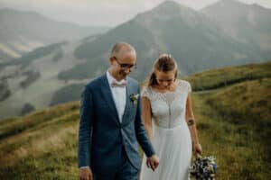 Ein Paar geht Hand in Hand einen Grasweg entlang, im Hintergrund eine Berglandschaft, und verkörpert damit die für Heiraten typische Essenz von Freude und Intimität. Der Mann trägt einen blauen Anzug und eine Fliege, während die Frau ein weißes Kleid trägt und einen Blumenstrauß hält, was die Magie ihrer Hochzeit einfängt.