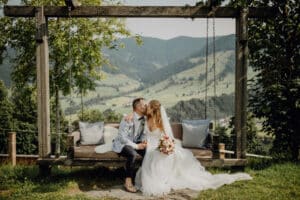 Ein frisch vermähltes Paar küsst sich auf einer Holzschaukel, umgeben von üppigem Grün und sanften Hügeln. Die Braut genießt die Ruhe ihrer Hochzeit, hält einen Blumenstrauß in der Hand und beide sind in Hochzeitskleidung gekleidet, was eine bezaubernde und romantische Szene schafft.