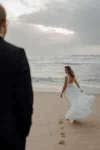 Eine Frau in einem fließenden weißen Kleid, das die Essenz einer Hochzeit verkörpert, läuft barfuß an einem Sandstrand entlang und hinterlässt Fußspuren. Sie blickt zurück auf einen Mann im Anzug, der im Vordergrund steht. Die Meereswellen und der bewölkte Himmel bilden eine ruhige Kulisse für diesen magischen Moment.
