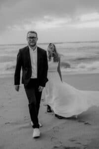 Ein Schwarzweißfoto zeigt ein Paar, das am Strand spazieren geht und sich vielleicht seine Hochzeit vorstellt. Der Mann trägt Anzug und Brille, während die Frau ein wallendes Kleid und Stiefel trägt, ihr Haar hält und vor dem Hintergrund der Meereswellen lächelt.
