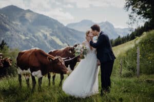 Braut und Bräutigam küssen sich in einer üppigen Berglandschaft, umgeben von braunen und weißen Kühen. Als Teil ihrer Hochzeit hält die Braut einen Blumenstrauß auf dem grasbewachsenen Hügel mit fernen Gipfeln unter einem blauen Himmel.