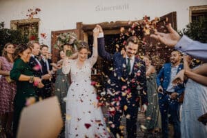 Braut und Bräutigam verlassen freudig den Veranstaltungsort und feiern ihre Hochzeit, während die Gäste mit Blütenblättern werfen. Die Braut hält einen Blumenstrauß in einer Hand, während der Bräutigam seinen triumphierend in die Höhe reckt. Sie sind von lächelnden Freunden und Familienmitgliedern umgeben, die alle festlich gekleidet sind und einen perfekten Hochzeitstag feiern.