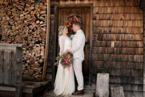 Ein Paar im Hochzeitskleid steht einander lächelnd gegenüber vor einem rustikalen Holzgebäude. Die Braut hält einen farbenfrohen Blumenstrauß in der Hand und beide sind in Weiß gekleidet. Ein Stapel Brennholz trägt zur gemütlichen Atmosphäre bei – ein bildschöner Moment für jeden, der in die Hochzeitsplanung vertieft ist.