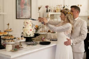 Inmitten einer wunderschön inszenierten Hochzeit schneiden Braut und Bräutigam elegant eine dreistöckige, mit Blumen geschmückte Torte an. Die Braut in ihrer Blumenkrone und ihrem Spitzenkleid steht neben dem Bräutigam in seinem beigen Anzug. Der Tisch ist übervoll mit Desserts und Dekorationen und zeugt von einer sorgfältigen Hochzeitsplanung.