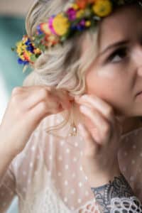 Eine Frau mit einem bunten Blumenkranz rückt ihren Perlenohrring zurecht und stellt sich dabei vielleicht ihre eigene Hochzeit vor. Sie trägt eine transparente, gepunktete weiße Bluse und zeigt ein detailreiches Tattoo auf ihrem Handgelenk. Ihr hellblondes Haar ist in lockeren Locken gestylt, was die Romantik der Hochzeitsplanungsträume noch verstärkt.
