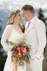 Braut und Bräutigam blicken sich lächelnd in die Augen, während sie ihre Hochzeit feiern. Die Braut trägt eine Blumenkrone und hält einen farbenfrohen Blumenstrauß, während der Bräutigam einen hellen Anzug trägt. Ein verschwommener natürlicher Hintergrund vervollständigt diesen bezaubernden Moment der Liebe und der tadellosen Hochzeitsplanung.