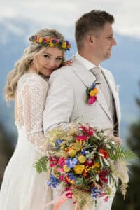 Eine Braut in einem Spitzenkleid und einer Blumenkrone umarmt einen Bräutigam in einem hellen Anzug und feiert ihre Hochzeit. Sie hält einen bunten Blumenstrauß mit blauen, gelben und rosa Blumen. Vor der atemberaubenden Bergkulisse blickt der Bräutigam zur Seite und verstärkt die Romantik ihres besonderen Tages im Freien.