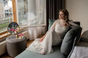 Eine Frau in einem weißen Spitzenkleid und einem Blumenstirnband sitzt auf einem Sofa am Fenster und träumt von ihrem Hochzeitstag. Auf einem kleinen Beistelltisch neben ihr stehen leuchtend rosa Blumen in einer Vase. Die Szene ist ruhig, weiches Tageslicht fällt durch das Fenster und schafft den perfekten Ton für die Hochzeitsplanung.