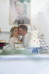 Braut und Bräutigam stehen liebevoll an ihrem Hochzeitstisch, der mit verschiedenen Torten geschmückt ist, darunter eine mehrstöckige Torte mit Blumendekorationen. Die Braut trägt eine Blumenkrone und ein Spitzenkleid, während der Bräutigam einen Anzug trägt. An der Wand hinter ihnen hängt ein skurriles Porträt.