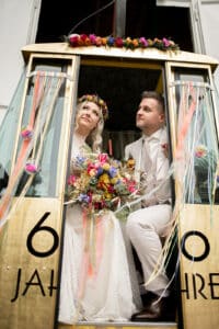 Ein Paar in Hochzeitskleidung sitzt in einer dekorierten Gondel, perfekt für ihre Hochzeit. Die Braut hält einen bunten Blumenstrauß und trägt einen passenden Blumenkranz. Die Gondel ist mit Luftschlangen und den Worten „60 Jahre“ an der Seite verziert.