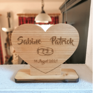 Ein Holzherz, perfekt für eine Hochzeit, zeigt die Namen „Sabine“ und „Patrick“ mit ineinander verschlungenen Ringen und dem Datum „14. August 2021“, elegant auf einem Holzständer platziert.