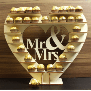 Ein herzförmiger Ständer präsentiert auf mehreren Ebenen elegant schokoladenüberzogene Leckereien, perfekt für eine Hochzeitsfeier. In der Mitte sind die Worte „Mr & Mrs“ in dekorativer Schrift ausgeschnitten, was jeder Hochzeitsplanung eine charmante Note verleiht.