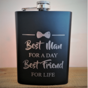 Auf einer schwarzen Flasche mit silbernem Deckel steht in weißen Buchstaben der Text: „Bester Mann für einen Tag, bester Freund fürs Leben“ mit einer Fliege darüber. Perfekt für die Hochzeitsplanung, steht sie auf einer Holzoberfläche vor einem hellen Hintergrund und zelebriert den Geist des Heiratens.