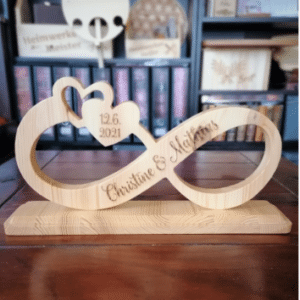 Eine hölzerne Skulptur mit Unendlichkeitssymbol, eingravierten Herzen und dem Datum „12.6.2021“, perfekt zur Erinnerung an Ihre Hochzeit. Sie trägt die Namen „Christine & Martin“ und steht elegant vor einem Bücherregalhintergrund.
