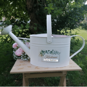 Eine weiße Gießkanne aus Metall, verziert mit Blumenmustern, steht auf einem Holztisch im Freien und verkörpert die Essenz einer romantischen Hochzeit. Eingravierte Unterschriften verstärken den Charme, während im Hintergrund zwischen einem Baum und üppigem Grün rosa Blumen blühen und die perfekte Kulisse für Hochzeiten schaffen.