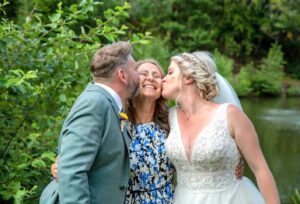 Eine Braut im weißen Kleid und ein Bräutigam im hellgrünen Anzug feiern ihre Hochzeit und küssen eine lächelnde Frau auf die Wangen. Sie stehen draußen neben einem Teich, mit üppigem Grün als perfekte Kulisse für ihren freudigen Anlass.