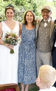 Eine Braut in einem weißen Kleid mit einem Blumenstrauß steht neben einer Frau in einem Kleid mit Leopardenmuster und einem Mann in einem grauen Anzug mit Schiebermütze. Sie stehen draußen inmitten üppiger Vegetation und lächeln herzlich. Der Kopf eines Kindes lugt in den Rahmen und fängt die freudige Essenz dieses wunderschönen Hochzeitsmoments ein.