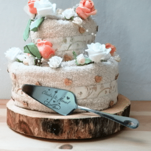 Zweistöckiger Handtuchkuchen mit weißen und pfirsichfarbenen Rosen auf einem Holzsockel, perfekt für alle, die schon lange mit der Hochzeitsplanung beschäftigt sind. Daneben sorgt ein Tortenheber mit Herz und Schriftzug für Charme. Ideal für eine einzigartige Hochzeits- oder Jubiläumspräsentation.