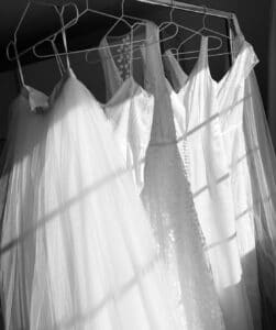 Eine Reihe weißer Brautkleider, die an einem Metallständer hängen, wartet auf die Magie der Hochzeitsplanung. Sonnenlicht fällt durch Jalousien und wirft gestreifte Schatten auf den Stoff. Die Kleider variieren in Textur und Design und erzeugen einen Kontrast zwischen glattem Satin und aufwendiger Spitze – perfekt für jede Hochzeit.