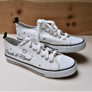 Weiße Sneaker mit schwarzen Akzenten, mit den Namen „Daniela & Marcel“ in eleganter Schrift und einem Herzdesign – perfekt für alle, die ihre Hochzeit planen. Diese hochzeitstauglichen Schuhe haben weiße Schnürsenkel und stehen auf einem Holzhintergrund.