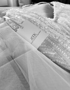 Nahaufnahme eines weißen, mit Perlen besetzten Kleides mit einem KTK-Etikett, perfekt zum Heiraten, mit zarten Spitzendetails und einem Schild mit der Aufschrift „KATHERINA MIKHAIL MOSCHEK“. Die komplizierte Textur des Stoffes unterstreicht das elegante Design.