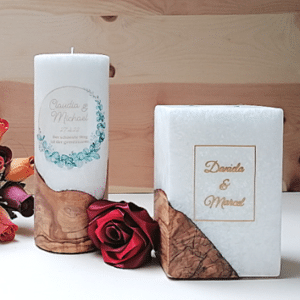 Zwei dekorative Kerzen, eine zylindrische und eine quadratische, sind mit Blumen- und Schriftmustern mit Namen versehen – perfekt für eine Hochzeitsfeier. Eine rote Rose und kleine orangefarbene Blumen stehen daneben vor einem Hintergrund mit Holzstruktur und schaffen eine elegante Kulisse für Ihre Hochzeitsplanung.