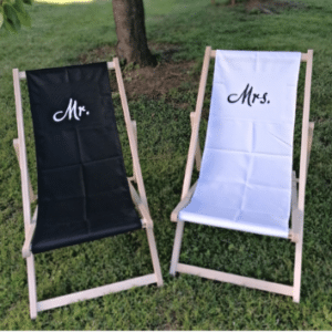 Zwei Holzliegestühle auf grünem Gras, einer mit schwarzem Stoffbezug und der Aufschrift „Mr.“ und der andere mit weißem Stoffbezug und der Aufschrift „Mrs.“ bilden die Bühne für eine perfekte Hochzeitsszene. Ein Baumstamm steht anmutig im Hintergrund und ergänzt diese idyllische Kulisse für diejenigen, die ihren Traumhochzeitsmoment planen.