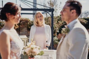 Ein Paar steht unter klarem Himmel vor einem Geistlichen und tauscht bei einer bezaubernden Hochzeit im Freien seine Gelübde aus. Die Braut, strahlend in ihrem weißen Kleid und mit dem Blumenstrauß in der Hand, steht neben dem Bräutigam, der in seinem hellen Anzug elegant aussieht. Ihre sorgfältig ausgearbeitete Hochzeitsplanung verleiht ihrem besonderen Tag Magie.
