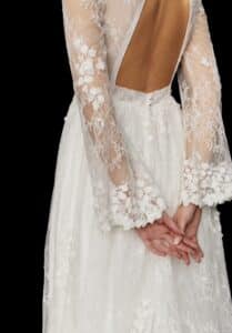 Eine Braut bei ihrer Hochzeit in einem Spitzenbrautkleid mit langen Ärmeln und offenem Rücken steht mit gefalteten Händen da. Das Kleid weist florale Details und komplizierte Spitzenmuster auf, perfekt für diejenigen, die sich in die Hochzeitsplanung vertiefen.