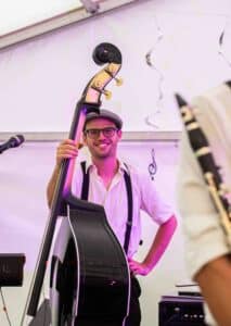Ein Musiker mit Brille und Schiebermütze lächelt und hält einen Kontrabass, was der Hochzeitsatmosphäre Charme verleiht. Lässig gekleidet mit Hosenträgern unter einem Zeltdach mit musikalischer Dekoration ergänzt er einen anderen, teilweise sichtbaren Musiker mit einer Klarinette.