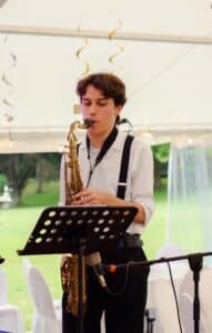 Ein junger Mann in weißem Hemd und schwarzen Hosenträgern spielt Saxophon in einem Zelt und verleiht der Hochzeitsatmosphäre Charme. Dekorative Saiten hängen von der Decke und draußen ist ein grüner Rasen zu sehen. Vor ihm auf einem Notenständer liegen Notenblätter, die den perfekten musikalischen Moment für die Heiratswilligen orchestrieren.