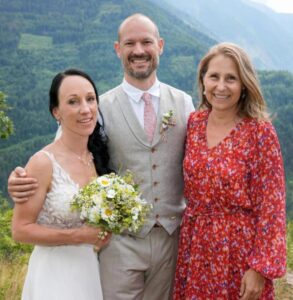 Eine Braut in einem weißen Kleid hält einen Blumenstrauß in der Hand. Sie steht neben einem Bräutigam in grauer Weste und Krawatte und einer Frau in einem roten Blumenkleid. Sie posieren draußen vor der üppig grünen Bergkulisse und fangen die Freude ihrer Hochzeit inmitten der Pracht der Natur ein.