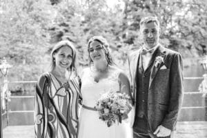Ein Schwarzweißfoto fängt einen Moment des Hochzeitsglücks ein, während drei Personen im Freien posieren. Eine Frau in einem gemusterten Kleid steht neben einer anderen Frau in einem weißen Kleid, die einen Blumenstrauß hält, während auf der anderen Seite ein Mann im Anzug steht. Bäume und ein See bilden die ruhige Kulisse.