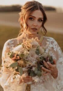 Eine Braut steht im Freien, versunken in die Freude ihrer Hochzeit, und hält einen Blumenstrauß in der Hand. Sie trägt ein Spitzenhochzeitskleid mit Blumenmuster und hat einen sanften Gesichtsausdruck. Der Hintergrund ist leicht verschwommen, wahrscheinlich ein Feld oder eine offene Fläche, mit warmem Licht, das darauf hindeutet, dass sie ihren perfekten Sonnenuntergang plant.